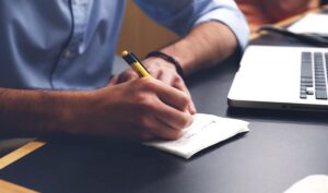 man reading from laptop and taking notes on piece of paper