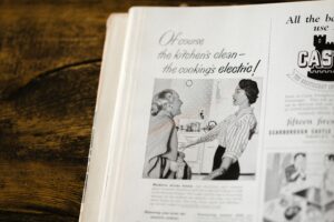Black and white print ad with two women in a kitchen. The younger one is telling the older one "Of course the kitchen's clean - the cooking's electric!"