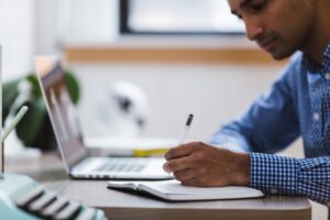 person writing on paper while sitting at desk with laptop