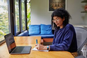 person writing on a piece of paper, sitting at desk with laptop
