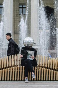 person with a mirrored ball on their head sitting on an outside bench reading a magazine entitled "Ideas"